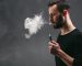 Young man vaping, studio shot. Bearded guy blowing a cloud of smoke on black background. Nicotine free smoking and vapor concept, copy space